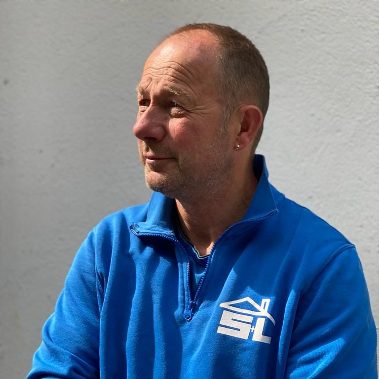 Mann mit kurzem Haar, trägt einen blauen Pullover mit Logo, schaut nachdenklich.