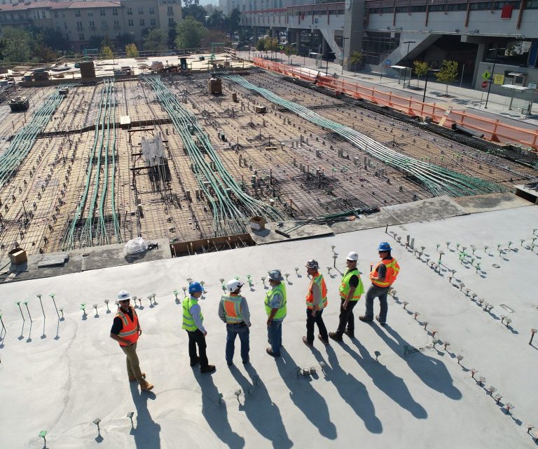 Arbeiter in Sicherheitskleidung auf einer Baustelle, umgeben von Rohren und Beton.